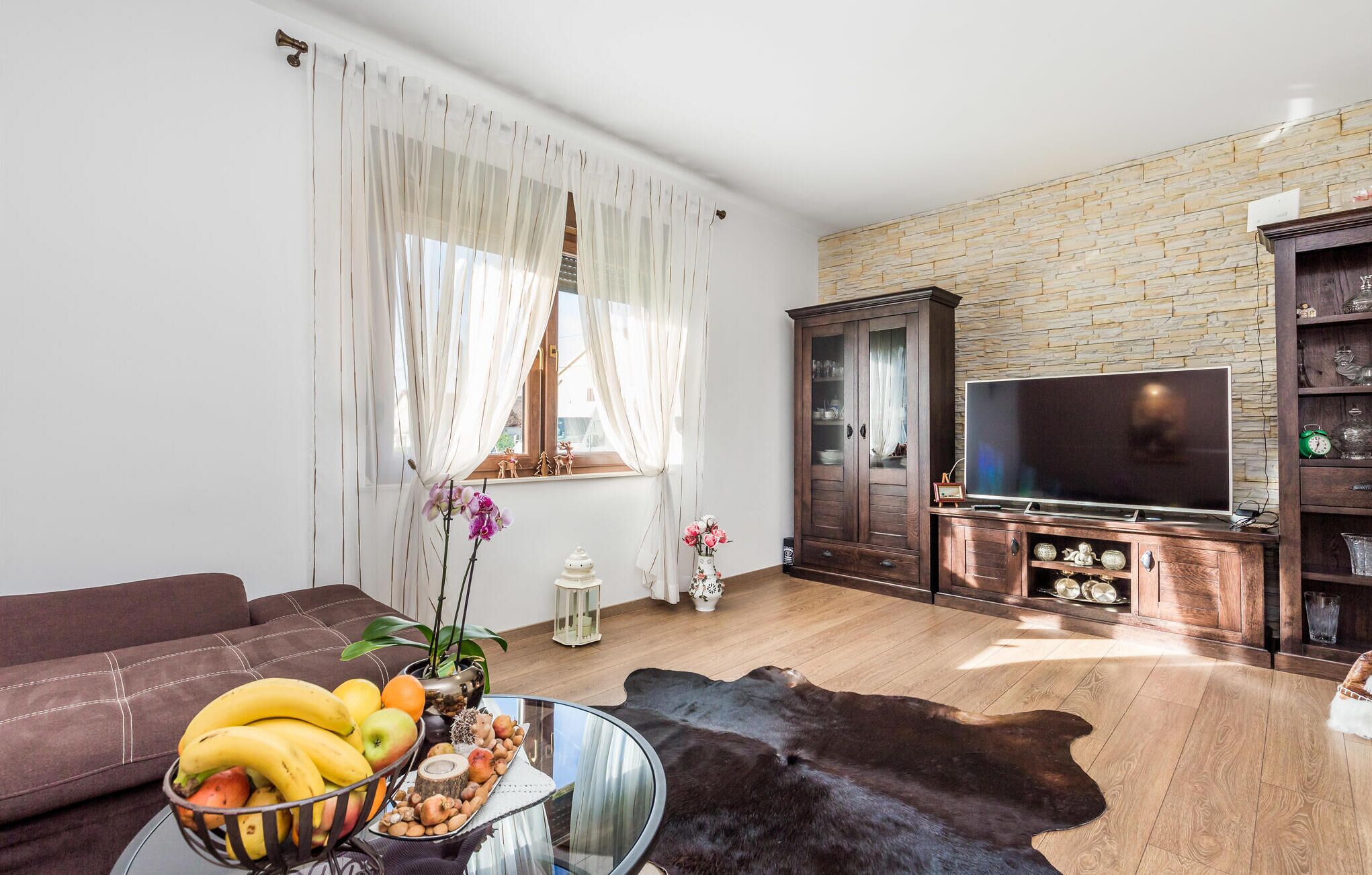 A living room with a sofa, a glass coffee table with fruit, a leather rug, a TV, and a stone wall.