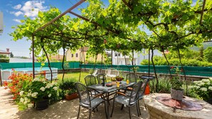 An outdoor patio with a table and chairs, surrounded by greenery and a pool in the background.