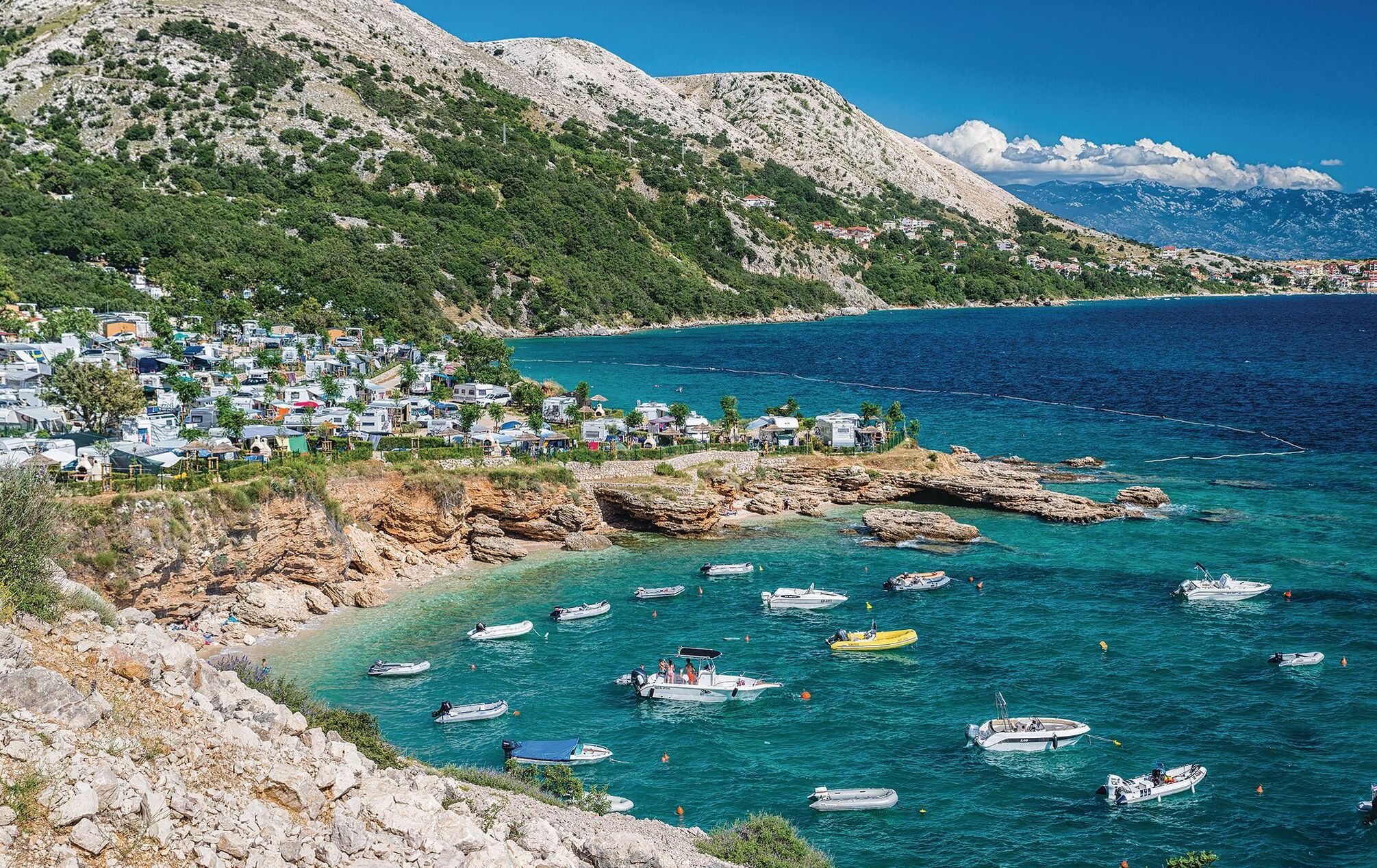 Une zone côtière avec un port de plaisance rempli de bateaux, une colline parsemée de bâtiments et, en arrière-plan, un paysage montagneux.