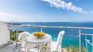 Un balcon avec une table et des chaises, un saladier rempli de fruits, et une vue sur la mer et le paysage lointain.