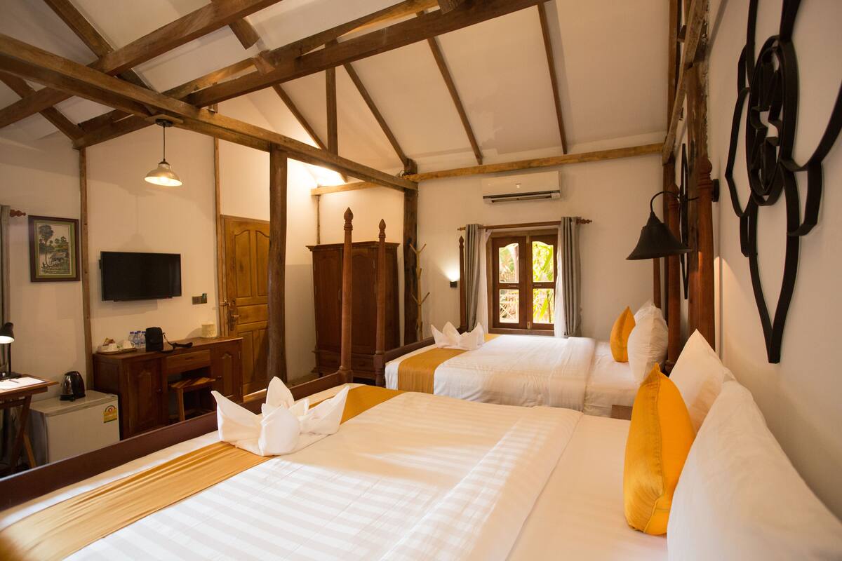 Une chambre d’hôtel avec deux lits, des poutres apparentes, une télévision, un bureau avec une bouilloire et une fenêtre avec des rideaux.