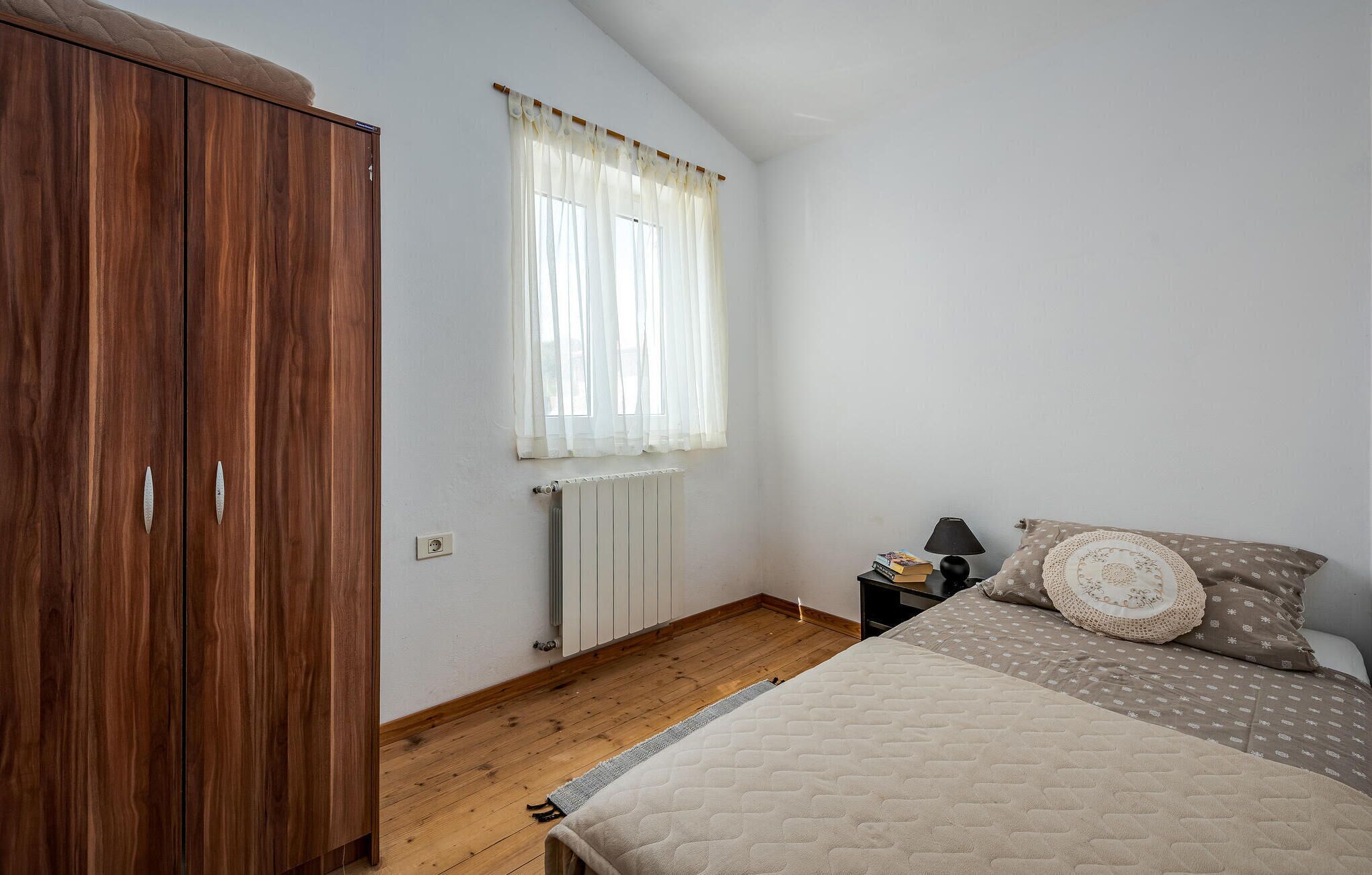 Quarto com guarda-roupa de madeira, cama com roupa de cama estampada, mesa de cabeceira com abajur e janela com cortinas finas.