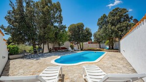 Una piscina con dos sillones blancos, rodeada de árboles y una pared blanca.
