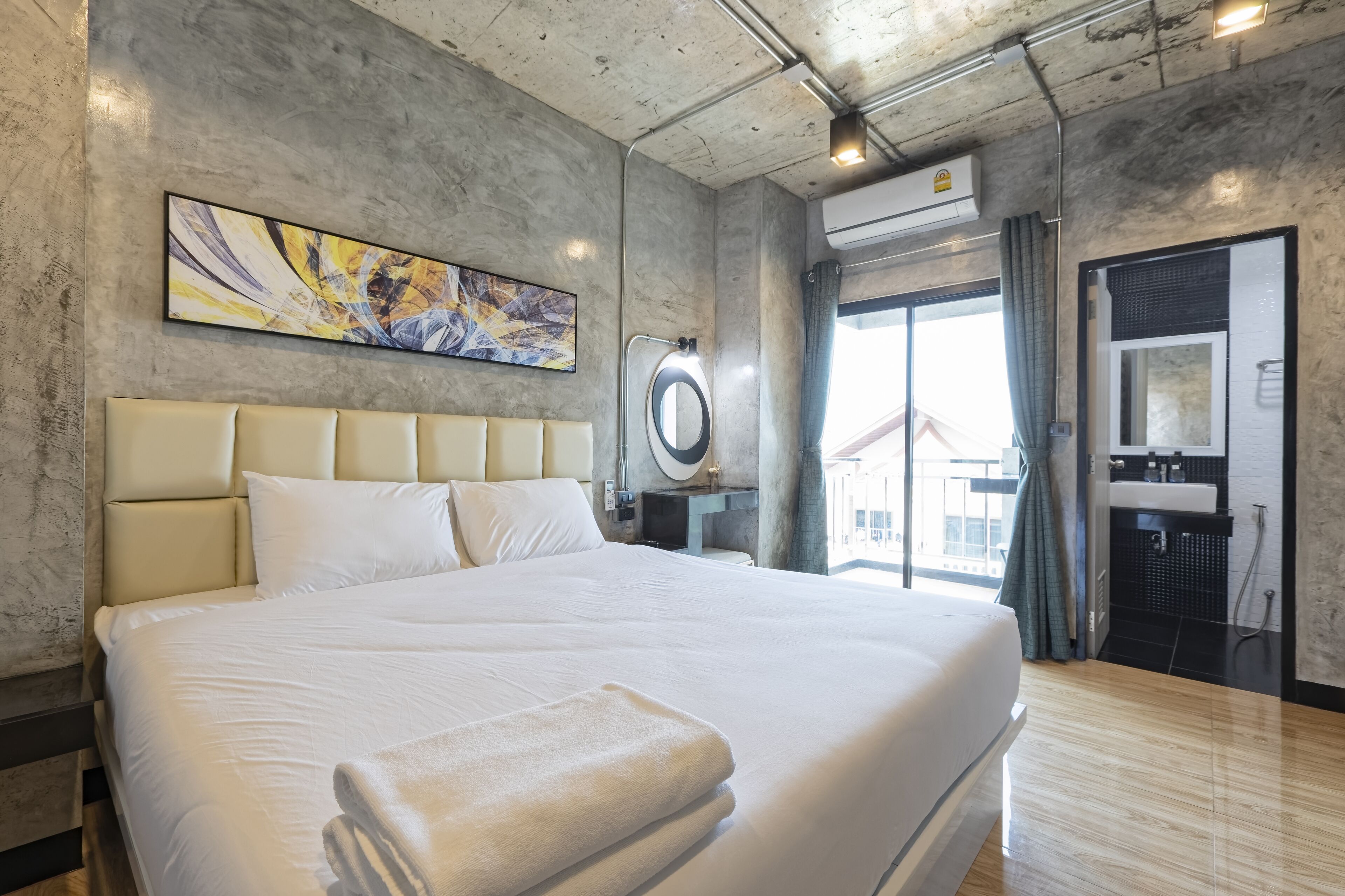 A modern hotel room with a large bed, a headboard, a painting, an air conditioning unit, a bathroom with a sink, and a view of the outside.