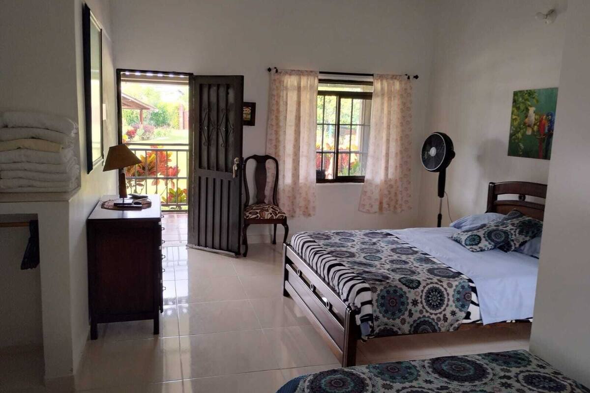 A bedroom with a bed, a chair, a desk with a lamp, a window with curtains, and a fan.