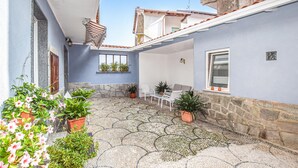 Un patio con suelo de baldosas, plantas en macetas, una zona de estar con sillas y una mesa, y un ventanal con persianas.