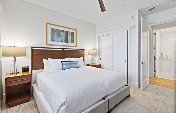 A neatly arranged bedroom with a large bed, two bedside tables with lamps, a framed picture on the wall, and a door leading to another room.