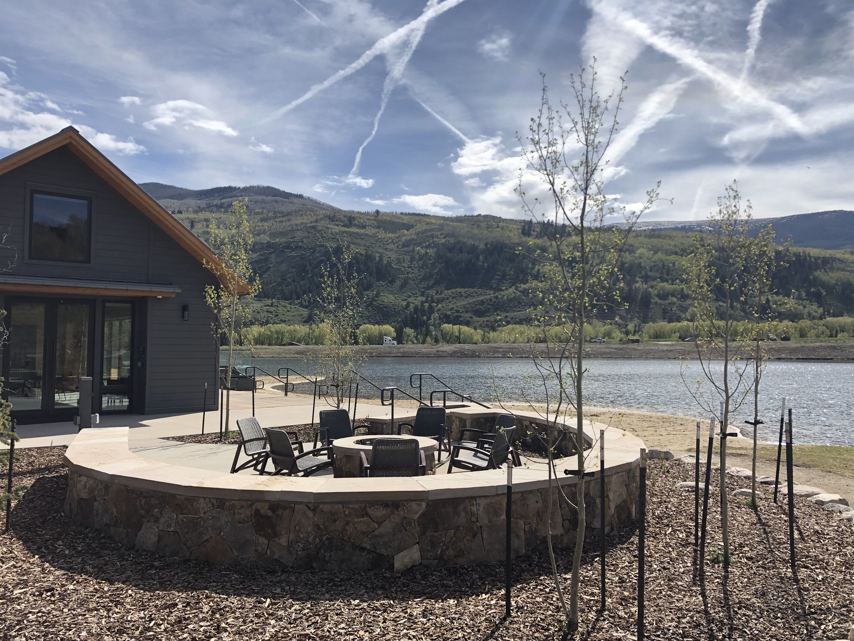 A lakeside patio with outdoor seating and a stone fire pit, surrounded by a stone wall and a view of a mountain and lake.