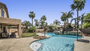 Piscina con cascada, rodeada de palmeras y un patio con mobiliario exterior.