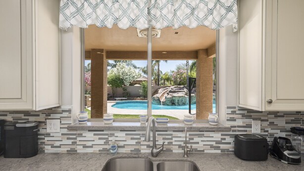Una cocina con un lavamanos doble, una ventana con vistas a un patio trasero con piscina y cascada, y un salpicadero de azulejos.