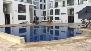 A modern indoor swimming pool with a tiled floor, surrounded by a multi-story building with balconies and large windows.
