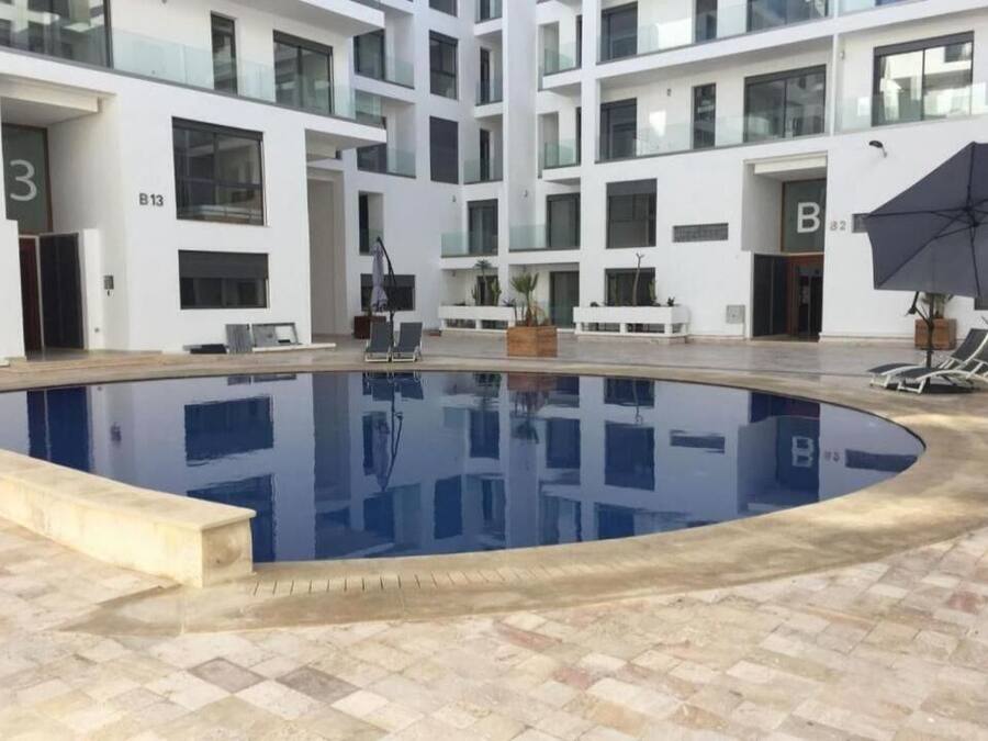 A modern indoor swimming pool with a tiled floor, surrounded by a multi-story building with balconies and large windows.