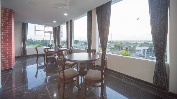 A dining area with a round table and chairs, large windows with curtains, and a view of the cityscape. - Bay Inn (Tirukalukundram)