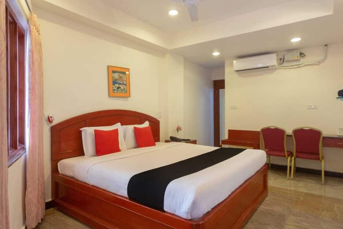 A hotel room with a wooden bed, red and white pillows, a desk with two chairs, and an air conditioning unit on the wall.