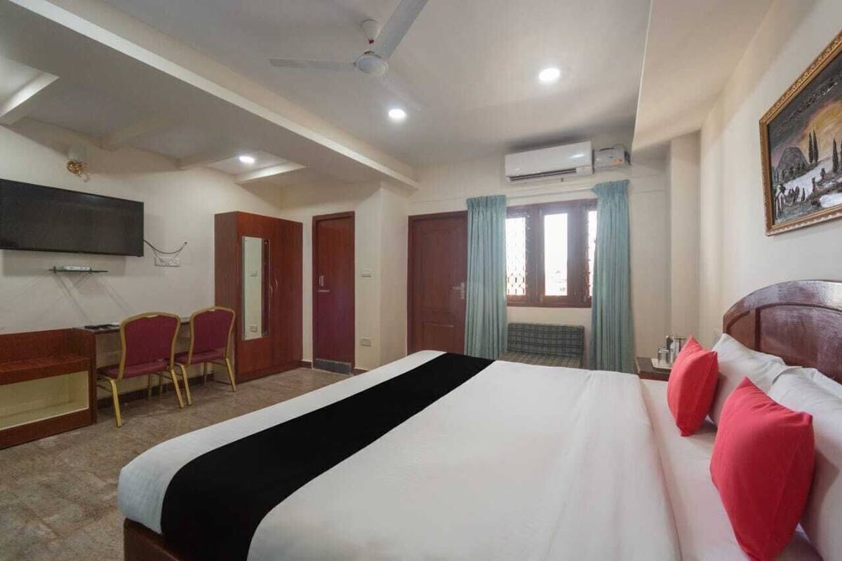A hotel room with a bed, a desk, chairs, a television, and an air conditioning unit.