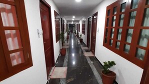 A long, narrow hallway with white walls and dark marble flooring. There are multiple doors with glass panels on both sides, and several potted plants are placed along the hallway. - Bay Inn (Tirukalukundram)