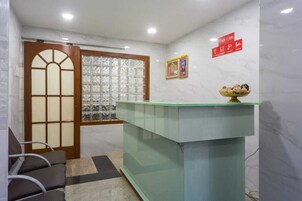 A reception area with a glass counter, a wooden door with glass panels, a waiting area with chairs, and a wall with framed pictures and a red sign.