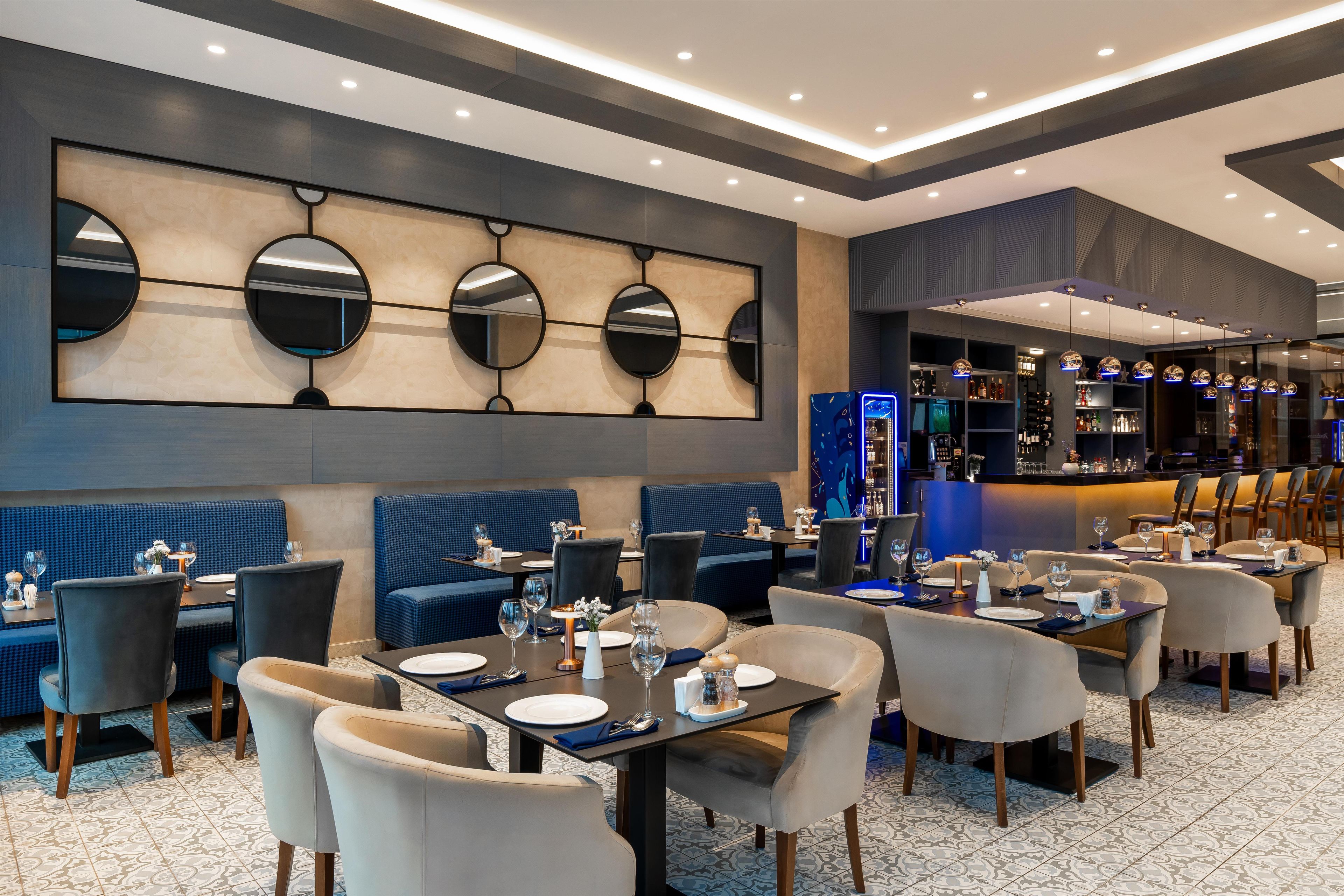 Un restaurante con asientos tapizados en azul, mesas redondas y una zona de bar con un letrero de neón azul.