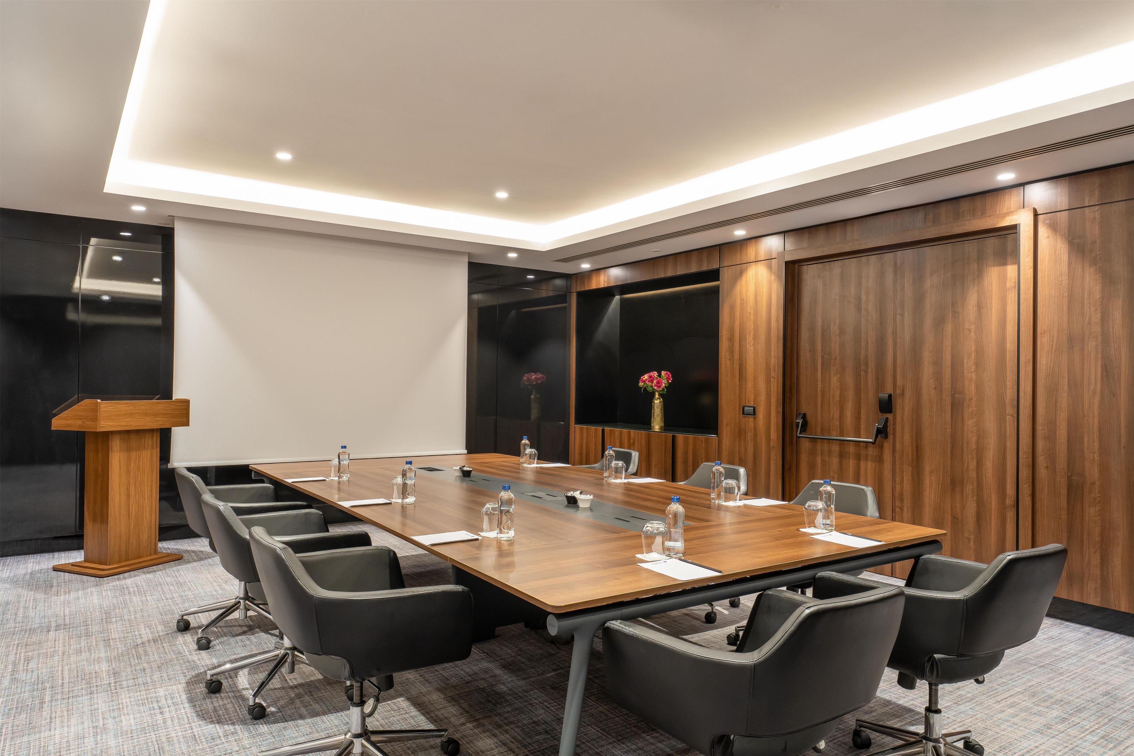 Une salle de conférence avec une longue table, des chaises noires, un pupitre et une grande armoire en bois.