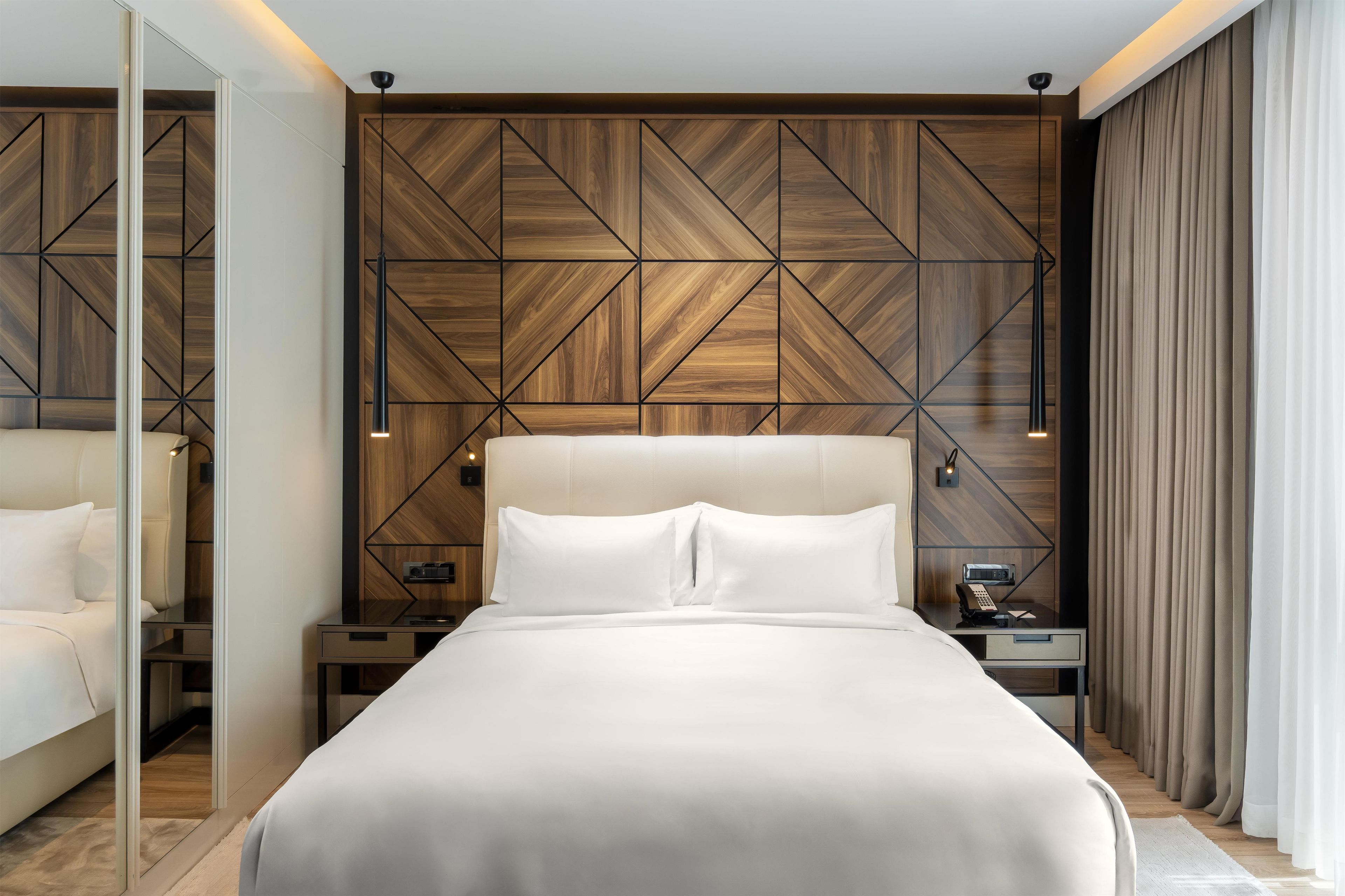 A modern bedroom with a geometric wood panel wall, a large bed with white bedding, bedside tables with lamps, and a mirror.