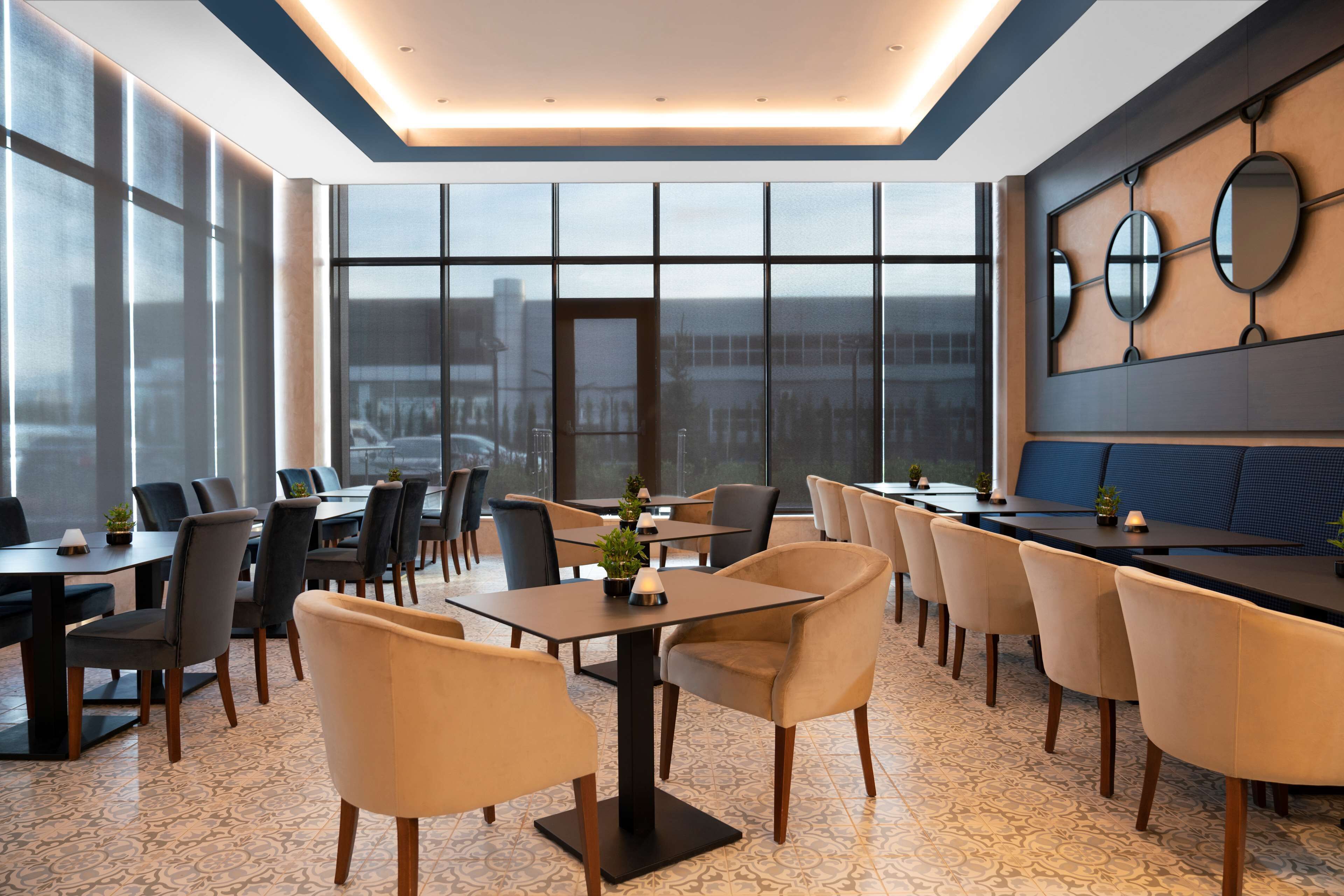 A modern dining area with multiple tables and chairs, large windows offering a view of the city, and decorative wall mirrors.