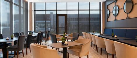 A modern dining area with multiple tables and chairs, large windows offering a view of the city, and decorative wall mirrors.