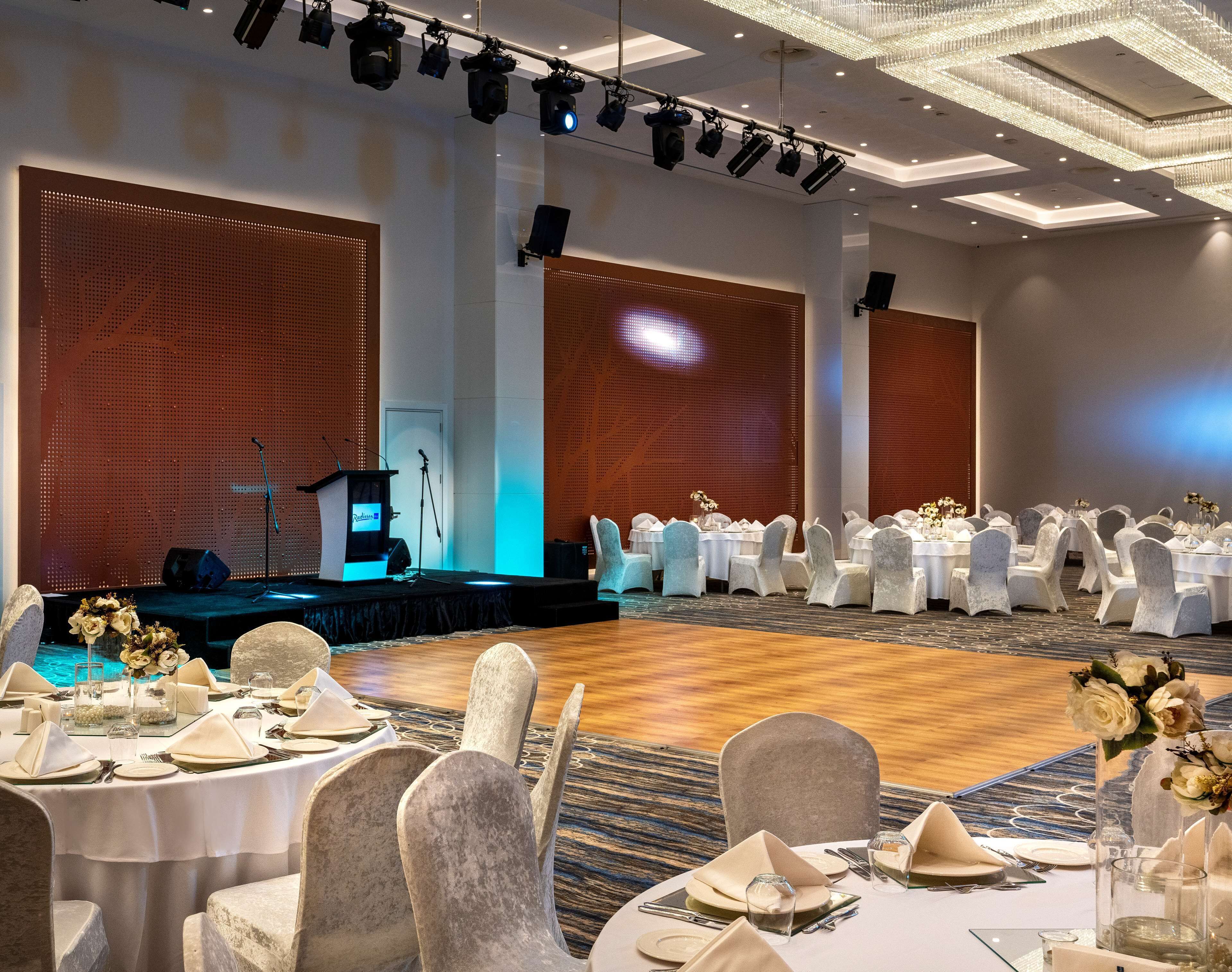 Een balzaal met ronde tafels gedekt voor een formeel evenement, een podium met lichtapparatuur en een groot scherm.