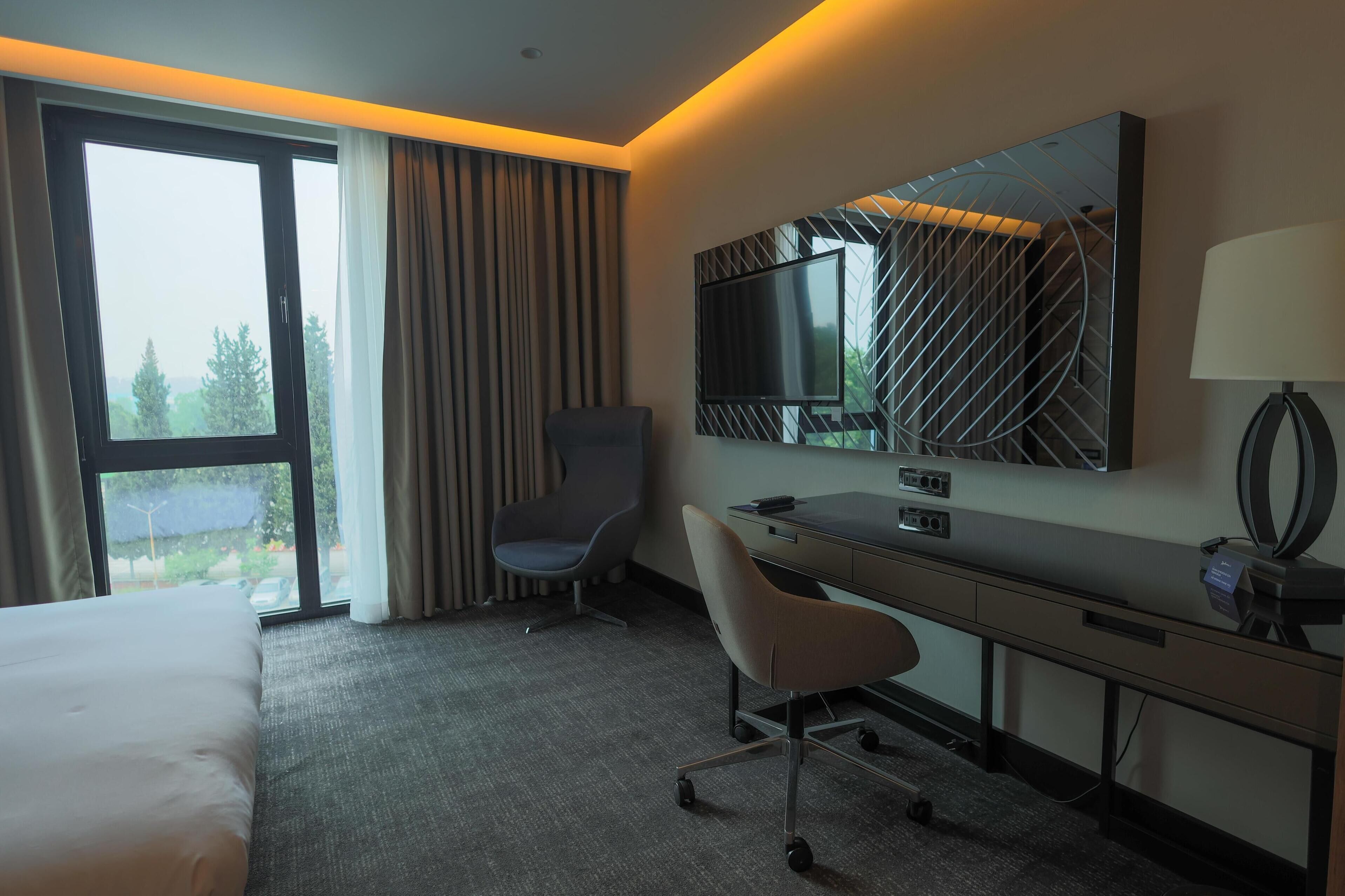 A hotel room with a bed, a desk, a chair, a TV, and a window with curtains.