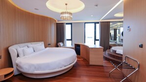 A modern hotel room with a curved bed, a desk, a chair, a small table, a hanging towel rack, and a large mirror. - Radisson Blu Hotel, Sakarya (Arifiye)