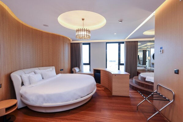 Una habitación de hotel moderna con una cama curva, un escritorio, una silla, una mesita pequeña, un toallero colgante y un espejo grande.