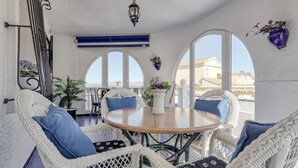 Una stanza luminosa con un tavolo rotondo in legno circondato da poltrone in vimini con cuscini blu, una parete bianca con persiana blu e una pianta in vaso sul davanzale.