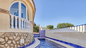 Una zona piscina stretta con pareti in pietra e piastrelle blu, che conduce a un edificio con un'ampia finestra ad arco e un balcone con ringhiere bianche.
