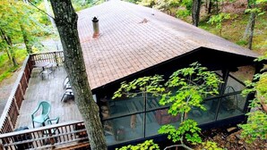 Una cabaña con terraza de madera, cerramiento de vidrio y vistas al bosque circundante.