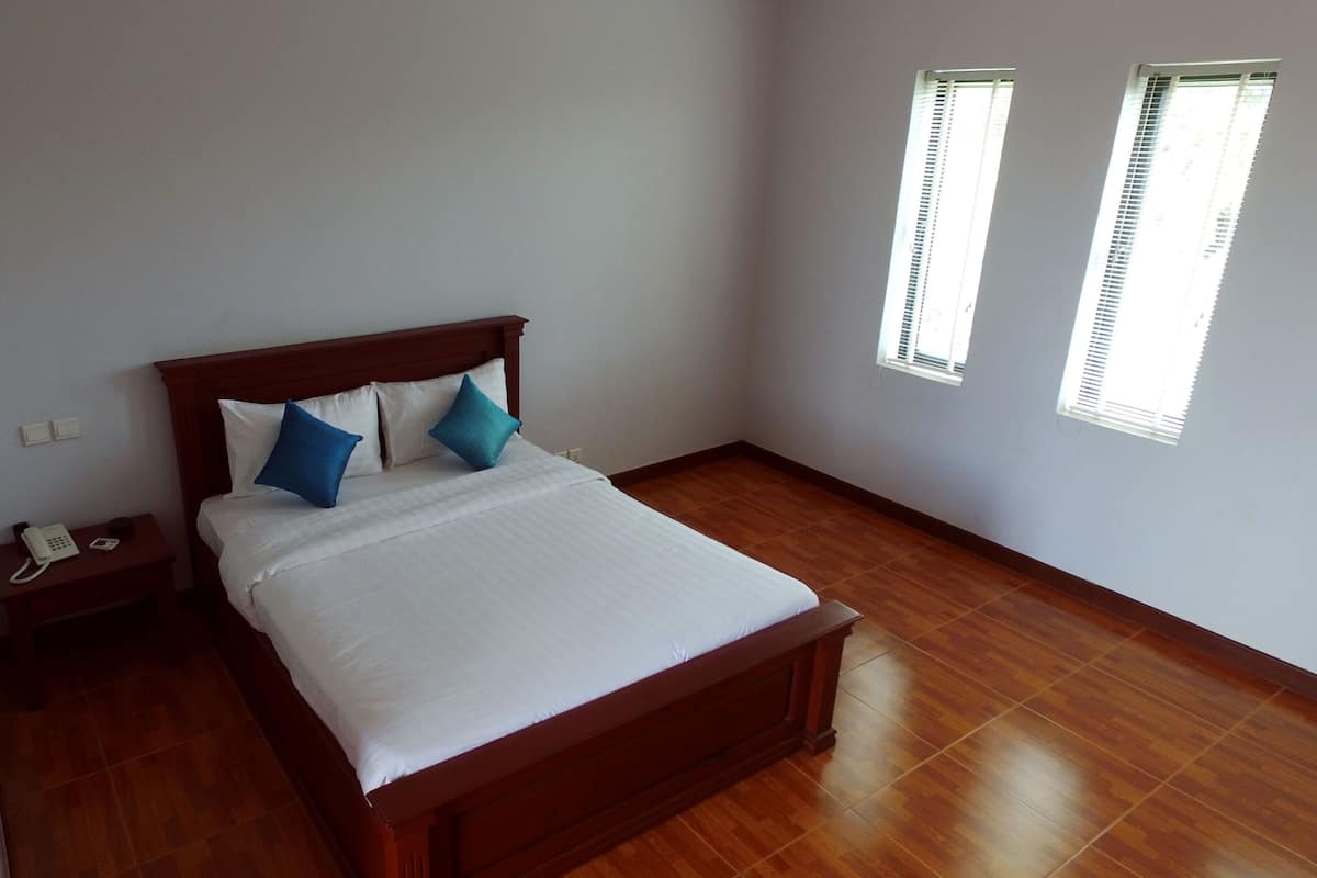 A neatly made bed with white bedding and two pillows, a wooden nightstand with a phone, and two windows with blinds.