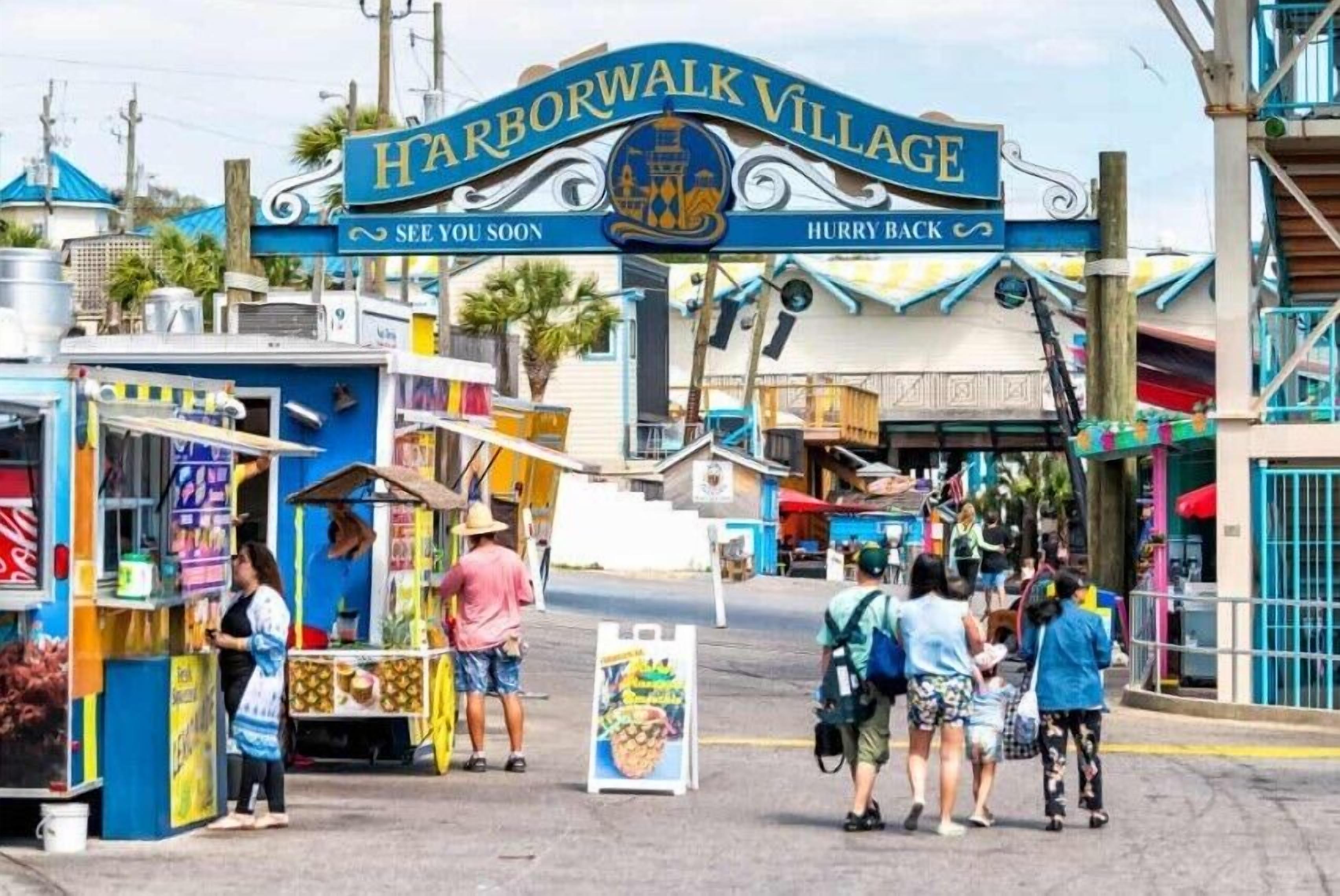 A colorful street scene with a sign reading "Harborwalk Village" and a small kiosk with a person standing nearby.
