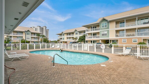A swimming pool with a brick deck and white lounge chairs, surrounded by a white fence.