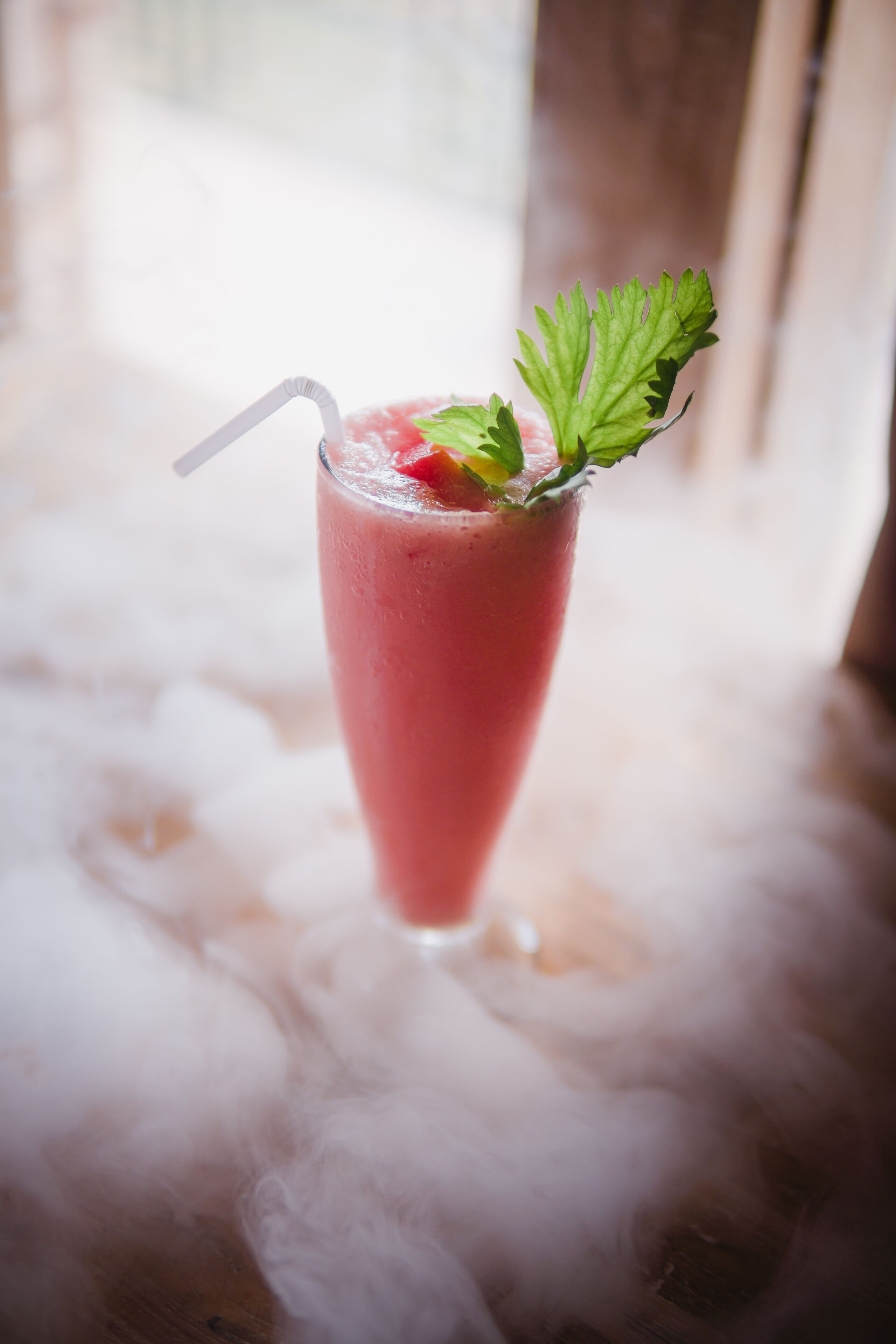 A tall glass of pink beverage with a straw and a mint garnish on a wooden surface.