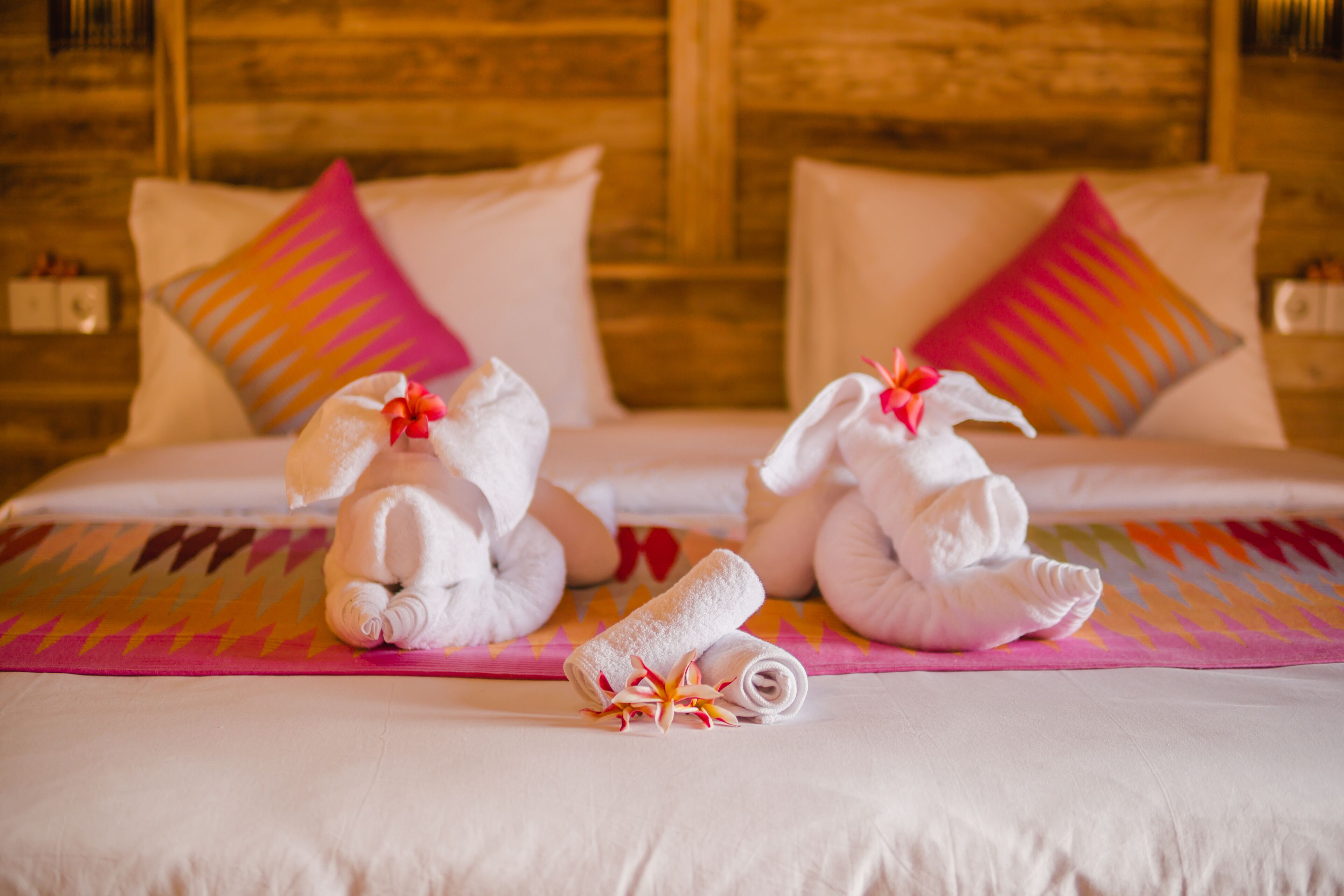 A hotel room with a bed featuring two rolled towels with flowers, a colorful bedspread, and wooden headboard.