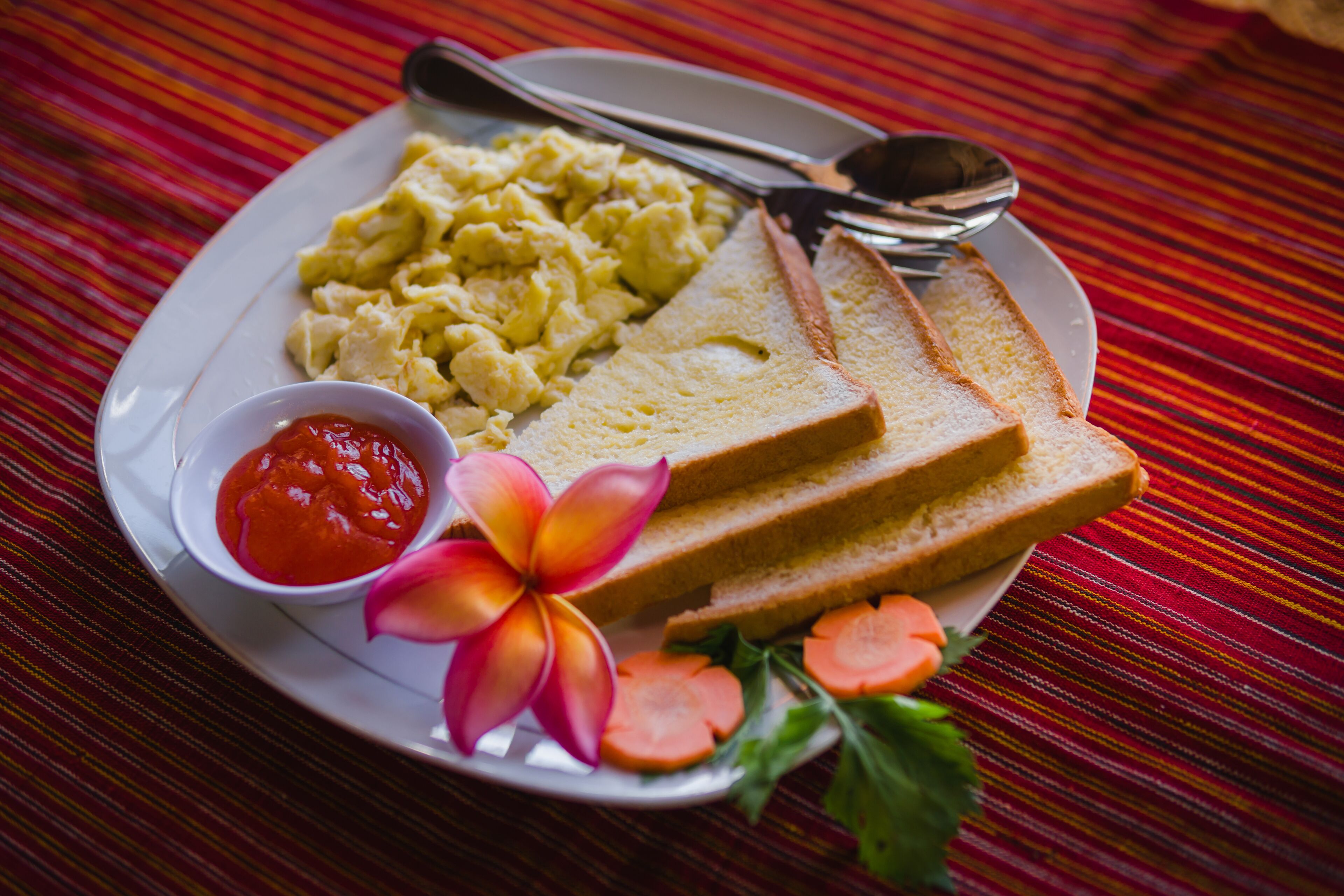 Un plato con huevos revueltos, tostadas, un recipiente pequeño de kétchup, una flor y algunas rodajas de zanahoria sobre un mantel a rayas.