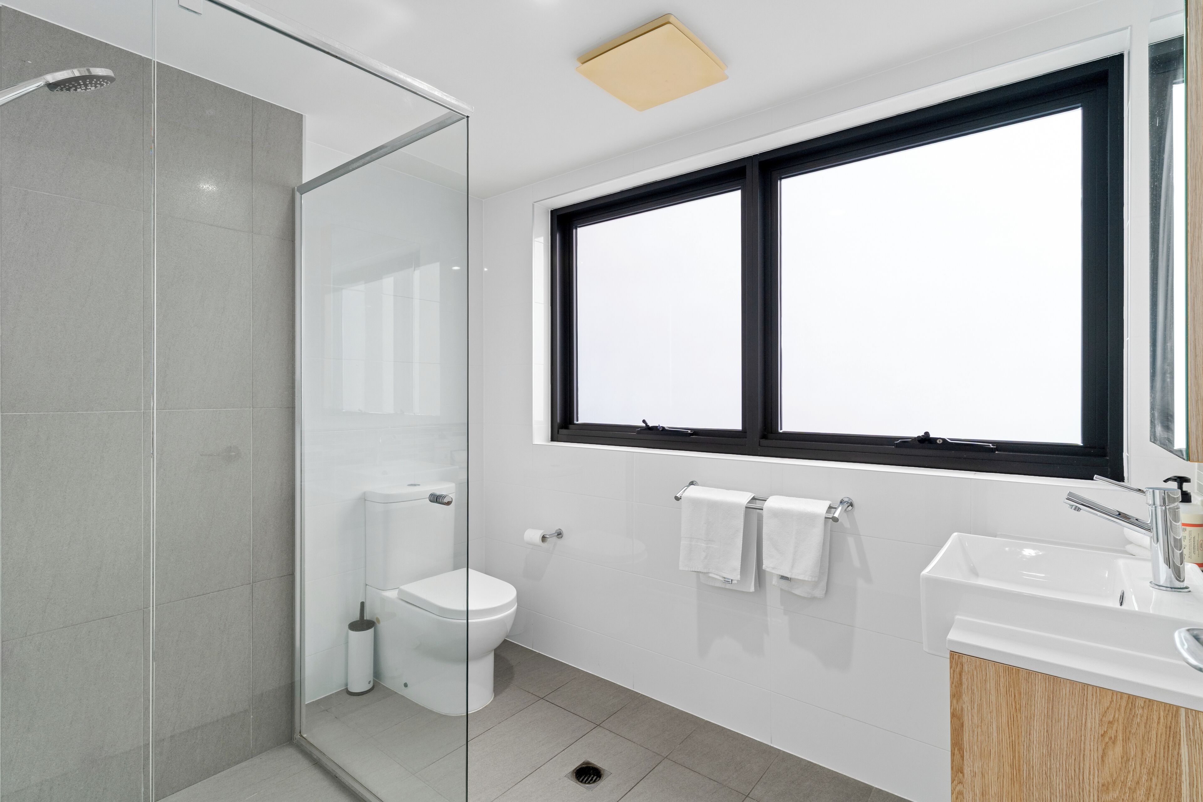 A modern bathroom with a glass shower enclosure, a toilet, a sink, and a window with a black frame.