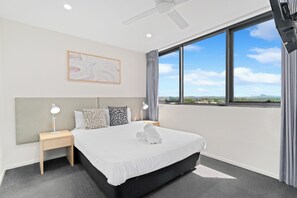 A modern hotel room with a large bed, a nightstand with a lamp, a ceiling fan, and a large window with a view. - Direct Collective - Rhythm on Beach (Maroochydore)