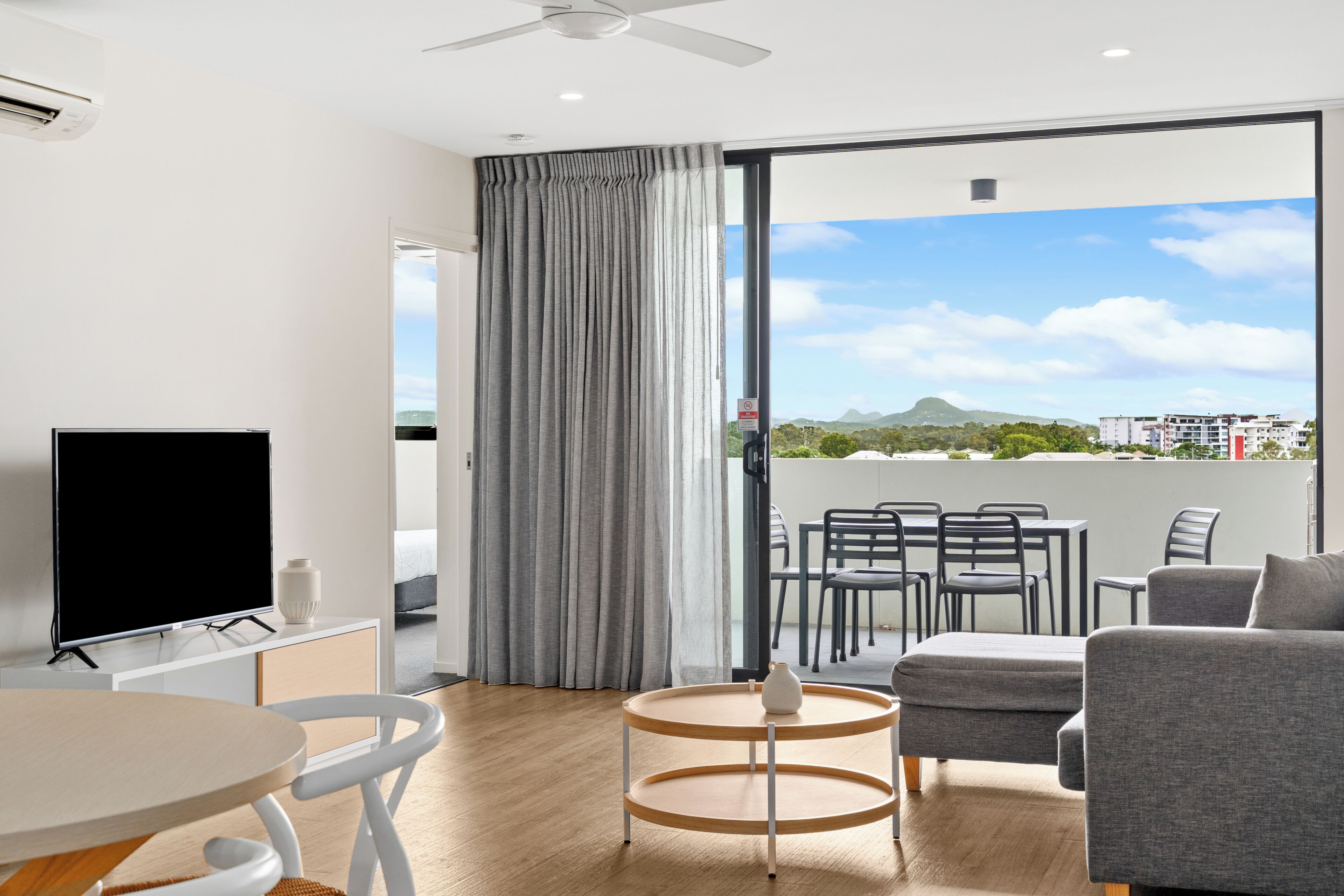 A modern room with a flat-screen TV, a dining table, a sofa, and a view of a balcony with outdoor furniture.