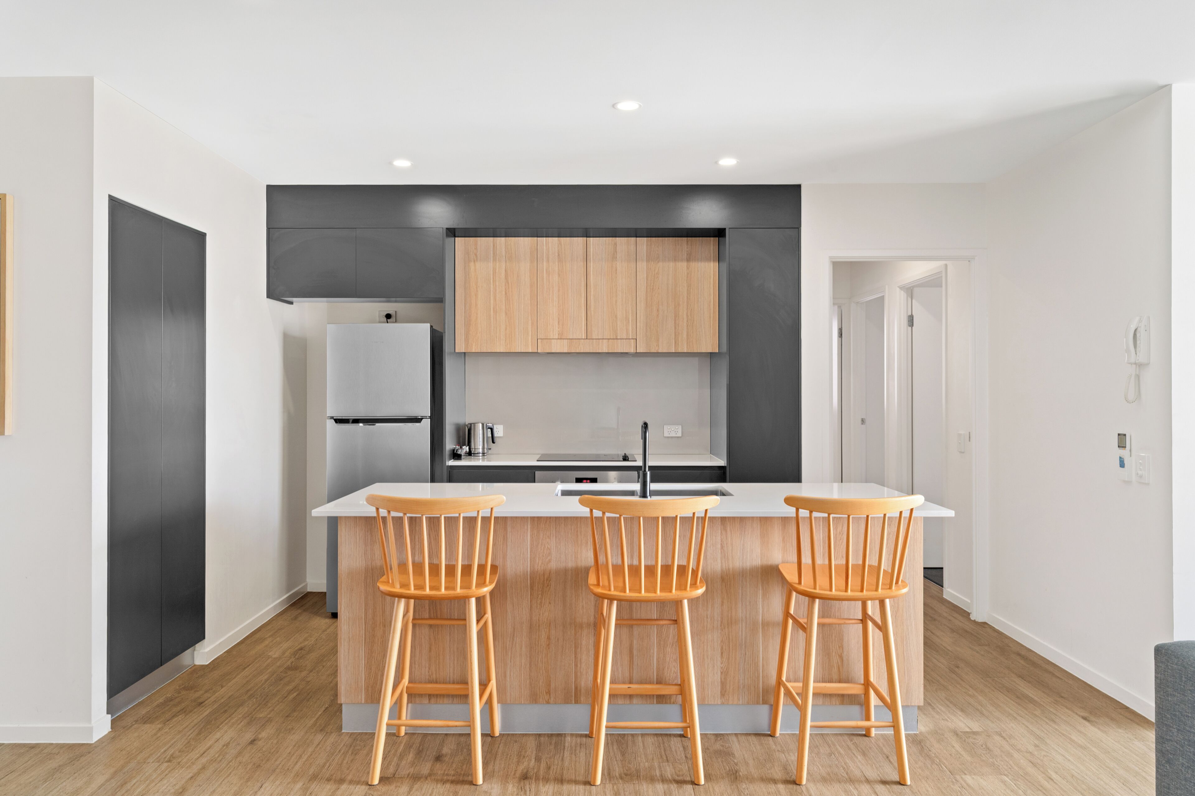 A modern kitchen with a central island, wooden bar stools, and a refrigerator.