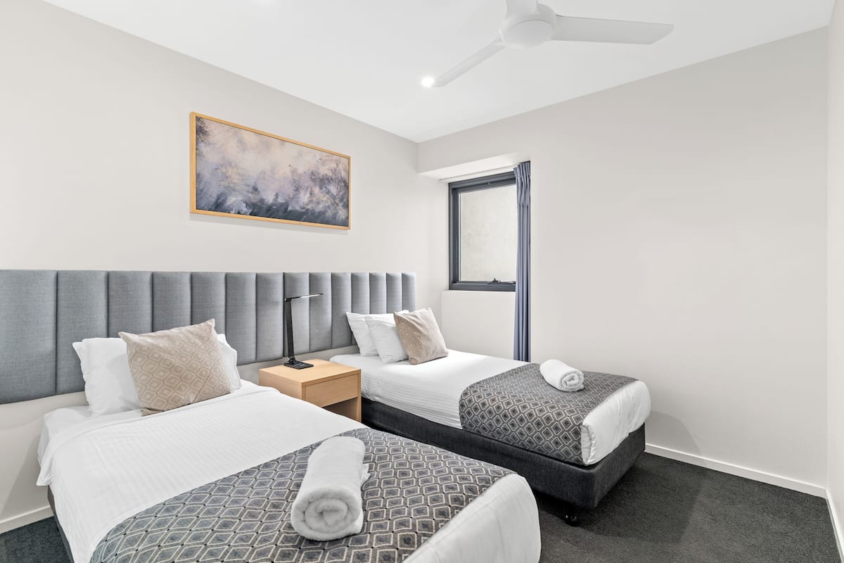 A hotel room with two beds, a ceiling fan, a window, and a painting on the wall.