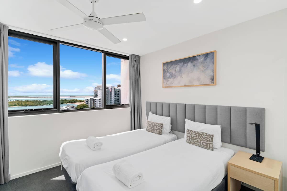 A hotel room with two beds, a ceiling fan, a large window with a city view, and a painting on the wall.