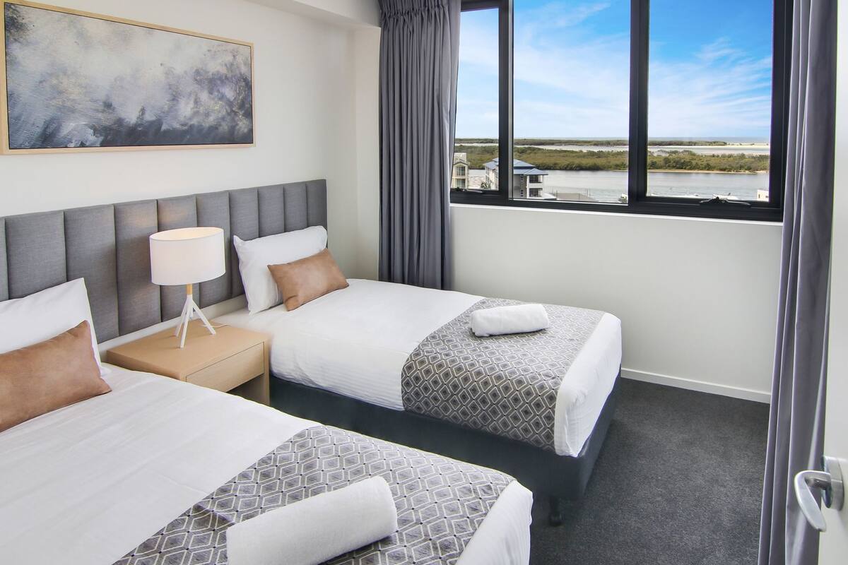 A hotel room with two beds, a nightstand, a lamp, a painting, and a window with a view of the beach and water.