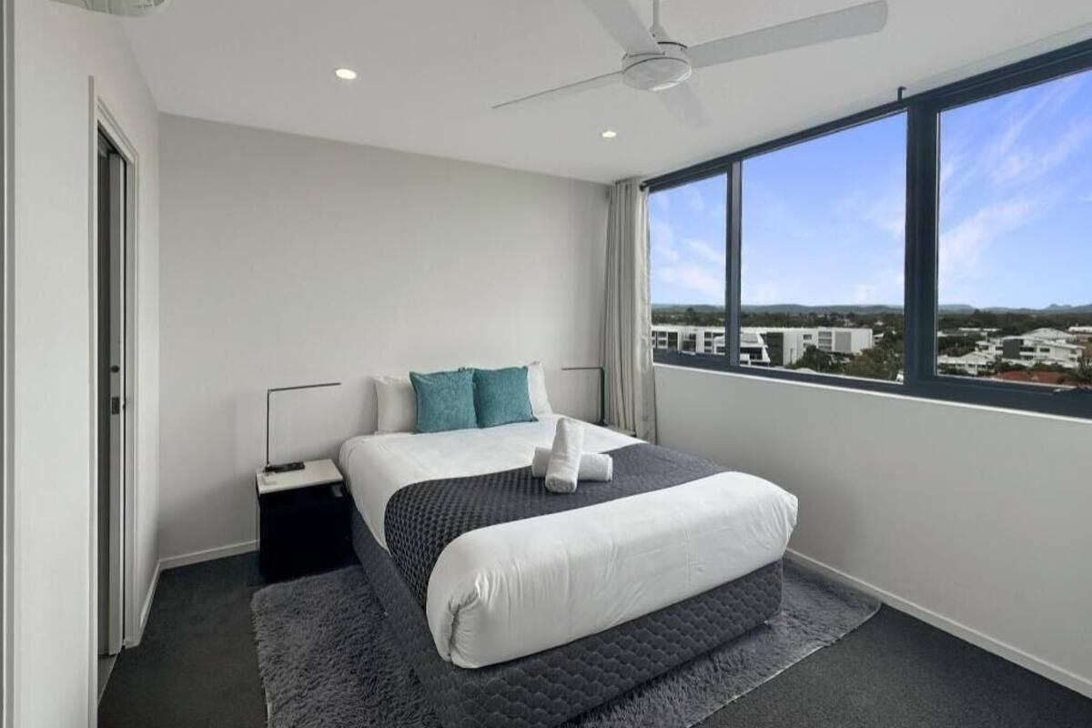A bedroom with a bed, a ceiling fan, a window with a view, and an air conditioning unit.
