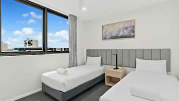 Une chambre d’hôtel avec deux lits, une tête de lit grise, une fenêtre avec des rideaux, un ventilateur de plafond et un tableau au mur.