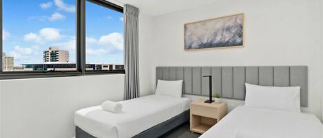 A hotel room with two beds, a grey headboard, a window with curtains, a ceiling fan, and a painting on the wall.