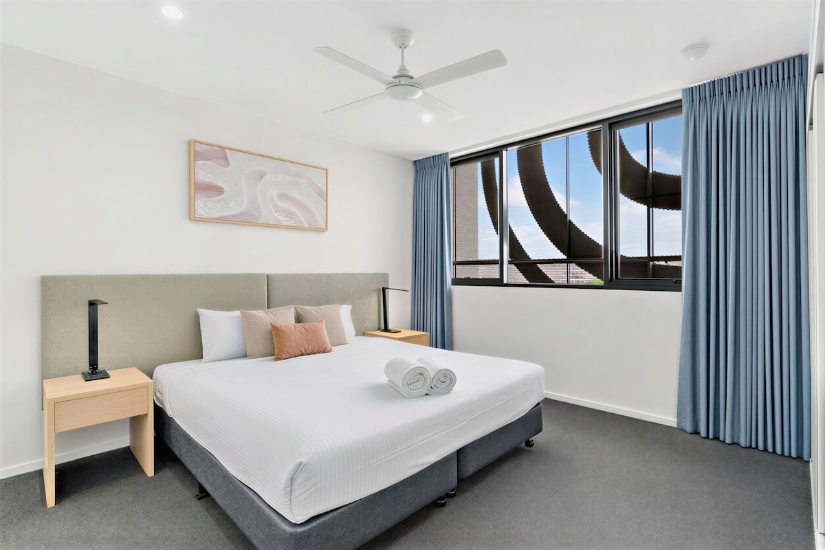 A modern hotel room with a large bed, a ceiling fan, a nightstand, a lamp, a framed picture, and a view of the sky through the window.